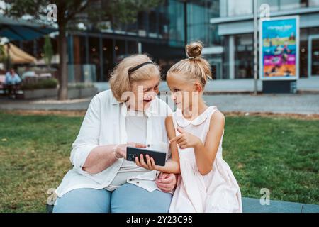 Oma holt das junge Mädchen von der Schule ab. Enkelin zeigt etwas auf dem Smartphone der Seniorengroßmutter. Stockfoto