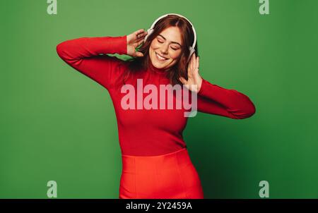 Glückliche junge Frau genießt die festlichen Feiertage, steht in einem grünen Hintergrund. Mit schnurlosen Kopfhörern und einem fröhlichen Lächeln streamt sie ihren Gefallen Stockfoto