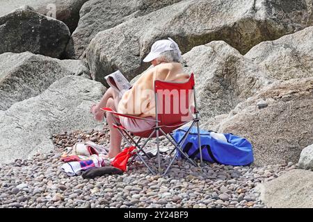 Seniorin Frau, die allein neben den Steinbrüchen am Kiesstrand sitzt, sonniger Tag, eingewickelt gegen die kühle Juni-Meeresbrise Sidmouth Devon England Großbritannien Stockfoto