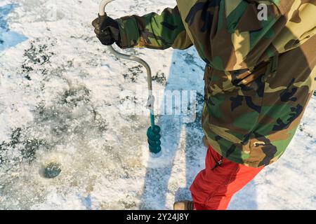 Ein warm gekleideter Asiate bohrt im Winter ein Loch ins Eis Stockfoto