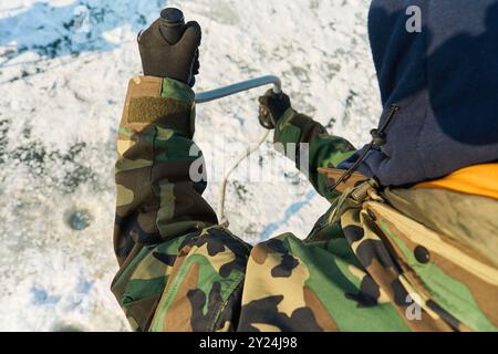 Ein warm gekleideter Asiate bohrt im Winter ein Loch ins Eis Stockfoto