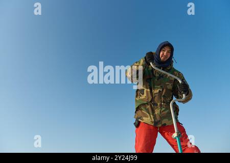 Ein warm gekleideter Asiate bohrt im Winter ein Loch ins Eis Stockfoto