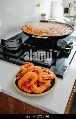 Nahaufnahme einer spanischen Fideua, einer Nudelpaella im Herd einer Hausküche und einem Teller mit frittierten Garnelen im Vordergrund Stockfoto