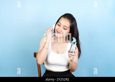 Glückliche asiatische Frau, die Baumwollkissen hält, Gesichtstuch oder mizellares Wasser auf ihr Gesicht aufträgt, Make-up entfernt und doppeltes Reinigungskonzept Stockfoto