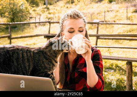 Die junge Frau entspannt sich mit ihrer Katze, während sie ferngesteuert an einem Laptop in einer sonnigen Umgebung auf dem Bauernhof arbeitet und die Natur und den Frieden genießt Stockfoto