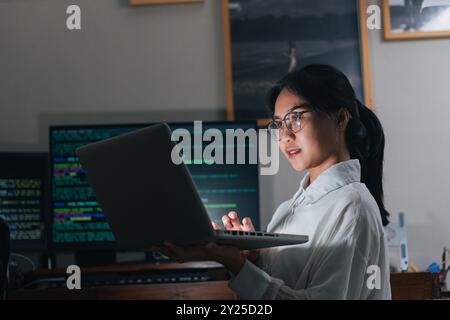 Eine junge Frau mit Brille arbeitet an einem Laptop in einem schwach beleuchteten Raum. Sie scheint konzentriert und engagiert zu sein, und ein Computerbildschirm zeigt den Code im an Stockfoto