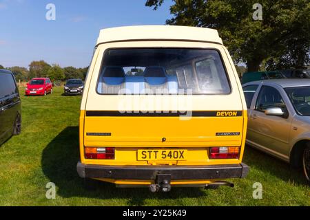 Rückansicht eines alten gelb-weißen VW-Transporters Y3 Wohnmobil, der auf einem grasbewachsenen Feld geparkt ist und von verschiedenen anderen Autos umgeben ist Stockfoto