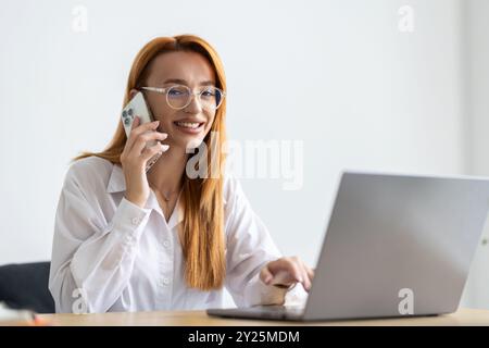 Bis morgen. Fröhliche junge, schöne Frau in Brille, die am Telefon spricht und mit einem Lächeln auf dem Laptop sitzt, während sie an ihrem Arbeitsplatz sitzt Stockfoto