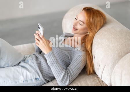 Lächelnde junge Frau, die ihr Handy benutzt, während sie zu Hause auf dem Sofa sitzt. Stockfoto