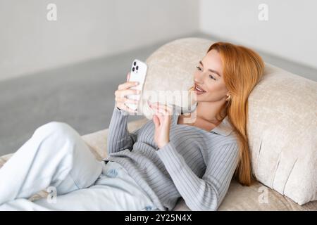 Lächelnde junge Frau, die ihr Handy benutzt, während sie zu Hause auf dem Sofa sitzt. Stockfoto