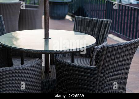 Ein runder Tisch mit Glasplatte, umgeben von vier Korbstühlen auf einer Terrasse. Die Umgebung ist im Freien mit einem verschwommenen Hintergrund von Grün. Stockfoto