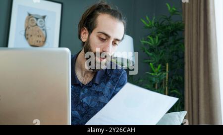 Ein Mann, der auf einer Couch sitzt, liest Dokumente, während er an seinem Laptop arbeitet. Er konzentriert sich zutiefst auf seine Aufgabe, indem er wichtige Papiere, Rechnungen oder FIN analysiert Stockfoto