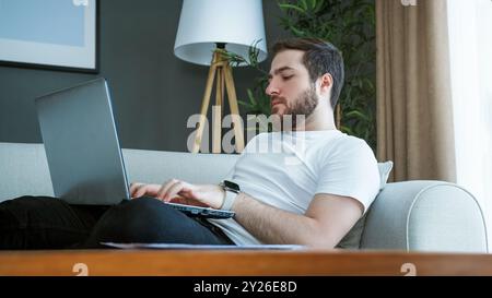 Ein Mann, der auf einer Couch liegt und an einem Laptop arbeitet. Konzentration und Verantwortung bei der Verwaltung persönlicher Finanzen, Rechnungen und wichtiger Papiere. Stockfoto