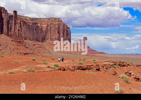 Zwei Wanderer auf dem Weg durch das Monument Valley mit weit entferntem Butte hinter dem östlichen Ende von Sentinel Mesa unter dramatischen Wolken Stockfoto