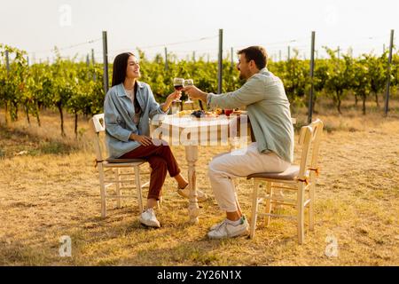 Ein paar entspannen sich an einem rustikalen Tisch in einem sonnigen Weinberg, genießen Wein und Gourmet-Snacks. Sie teilen Lachen und Freude, umgeben von üppigen Weinstöcken und dem Stockfoto