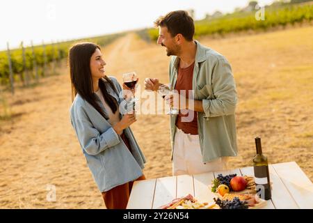 Ein paar entspannen sich an einem rustikalen Tisch in einem sonnigen Weinberg, genießen Wein und Gourmet-Snacks. Sie teilen Lachen und Freude, umgeben von üppigen Weinstöcken und dem Stockfoto