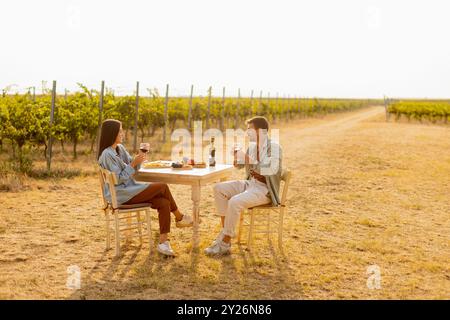 Ein paar entspannen sich an einem rustikalen Tisch in einem sonnigen Weinberg, genießen Wein und Gourmet-Snacks. Sie teilen Lachen und Freude, umgeben von üppigen Weinstöcken und dem Stockfoto