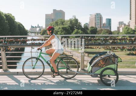 Eine Frau, die Fahrrad fährt, einen Anhänger zieht, mit einem Kind drin. Stockfoto