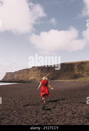 Frau in rotem Kleid, die auf schwarzem Sandstrand läuft, mit Klippen im Hintergrund Stockfoto
