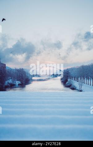 Schneebedeckte Hängebrücke im Rauch über einem ruhigen Fluss an einem wunderschönen Wintermorgen bei Sonnenaufgang Stockfoto