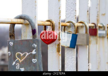 Nahaufnahme verschiedener Vorhängeschlösser, darunter ein rotes herzförmiges Schloss, das an einem rostigen weißen Metallgeländer befestigt ist, das Liebe, Engagement und Erinnerungen symbolisiert Stockfoto