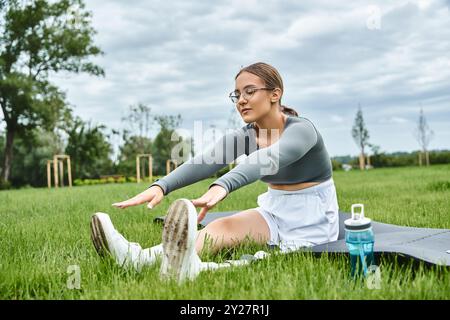 Eine junge Frau streckt ihre Beine, während sie an einem bewölkten Tag auf einer Matte in einem üppig grünen Park trainiert. Stockfoto