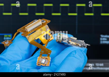 Optischer Sensor, Laserdiode, Steckverbinder oder Linse an flexiblen Kabeln vom CD-DVD-Laufwerk-Aufnahmekopf in der menschlichen Hand auf einem Labor-Oszilloskop-Bildschirm Detail. Stockfoto