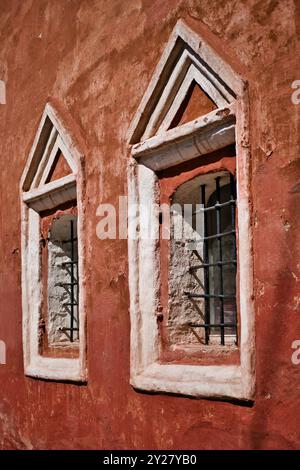 Zwei Fenster mit Metallkäfig in einer Reihe an der Wand eines alten roten Gebäudes. Altes Steingebäude. Einfacher dekorativer Rahmen um das Fenster. Stockfoto