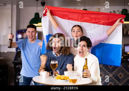 Eine Gruppe von begeisterten jungen erwachsenen Sportfans, die die niederländische Flagge schwenken und die Nationalmannschaft mit Bier im Pub unterstützen Stockfoto