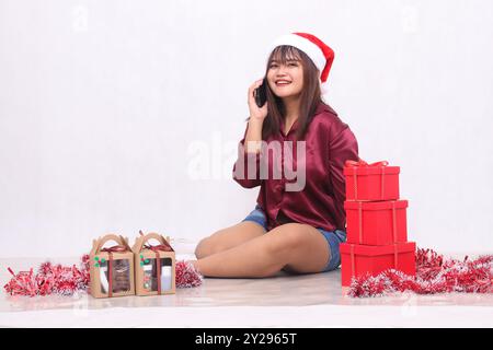 Fröhliches junges, schönes südostasiatisches Mädchen am Telefon neben 3 Geschenkboxen an Weihnachten, das Weihnachtsschmuck und rotes Hemd auf weißem Hintergrund trägt Stockfoto