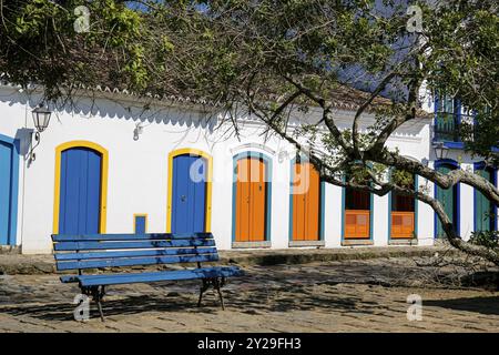 Blick auf die Fassaden des Kolonialhauses mit farbenfrohen Holztüren in der Sonne mit einer blauen Bank und Baumzweigen vor der historischen Stadt Paraty, Brasilien, Unes Stockfoto