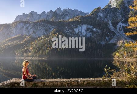 Eine Frau, die Yoga am See macht. Lotus-Position (Padmasana). Vorderer Gosausee im Herbst mit Blick auf das Dachsteingebirge. Der Gosaukamm auf dem Stockfoto