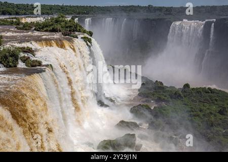 Blick auf die mächtigen Iguazu-Fälle in der Nähe von Devil's Throat mit braunem und weißem Wasser in üppiger grüner Vegetation Stockfoto