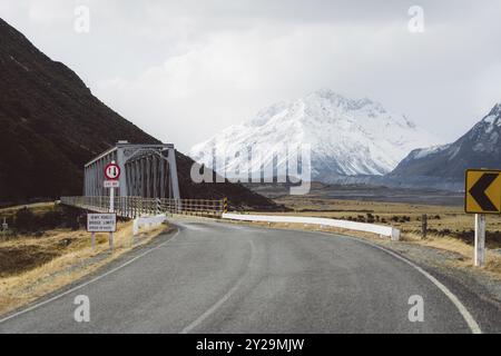 Straße, die zu einer Brücke mit schneebedeckten Bergen und bewölktem Himmel im Hintergrund führt, Hooker Valley Track, Neuseeland, Ozeanien Stockfoto