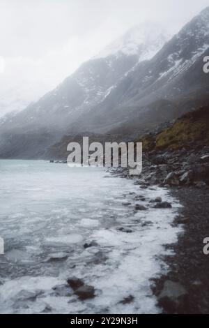 Blick auf einen gefrorenen See vor schneebedeckten Bergen mit nebeligem Himmel und felsiger Küste, Hooker Valley Track, Neuseeland, Ozeanien Stockfoto