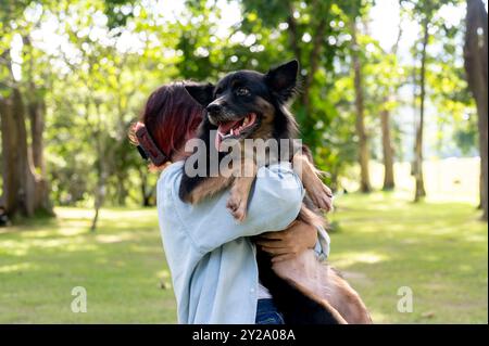 Eine Frau, die ihren Hund in den Armen hält, während sie durch einen grünen Park geht, ihr Haustier umarmt und gemeinsam einen lustigen Outdoor-Moment genießt. Tier, Tier, am besten Stockfoto