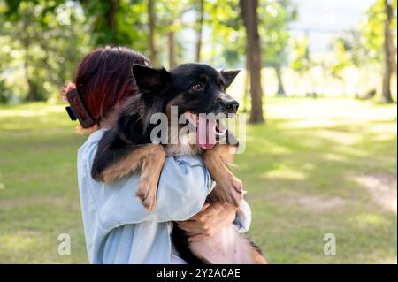 Eine Frau, die ihren Hund in den Armen hält, während sie durch einen grünen Park geht, ihr Haustier umarmt und gemeinsam einen lustigen Outdoor-Moment genießt. Tier, Tier, am besten Stockfoto