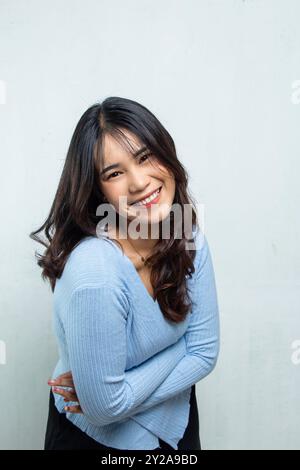 Nahaufnahme des asiatischen Schönheitsgesichts, das vor der Kamera posiert und ihren eigenen Körper umarmt, die Frau trägt ein blaues lässiges Top, lange fließende Haare und lächelt frei, ich Stockfoto