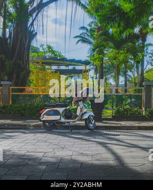 Balikpapan, Indonesien - 22. Juni 2024. Ein klassischer weißer Vespa-Roller, der auf einer Kopfsteinpflasterstraße geparkt ist, mit üppigen Palmen und einem lebhaften Garten in t Stockfoto