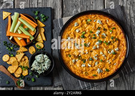 buffalo Chicken Dip in Backform auf grauen rustikalen Holztisch mit Crackern, Blaukäse, Karotten- und Selleriestangen auf Schieferbrett, horizontale Ansicht fr Stockfoto