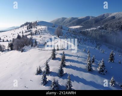 Blick aus der Vogelperspektive auf die Winterlandschaft mit schneebedeckten Hügeln am Morgen. Schneedecken und immergrüne Bäume. Klarer blauer Himmel steht im Kontrast zu weißem Schnee und ferne Berge bieten eine atemberaubende Kulisse. Stockfoto