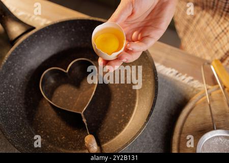Eine Person, die ein Ei mit einer gebrochenen Schale hält, bereit, es in einer Pfanne in eine herzförmige Form zu zerbrechen. Die Kücheneinrichtung ist warm und einladend Stockfoto