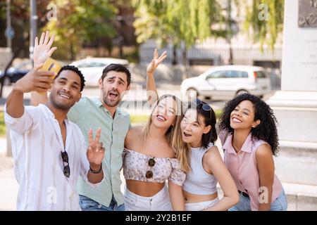 Fünf Freunde lächeln, winken und machen an einem sonnigen Tag ein lebhaftes Selfie im Freien, um einen lustigen Moment zusammen mit fröhlichen Ausdrücken und einer entspannten Atmosphäre festzuhalten. Stockfoto