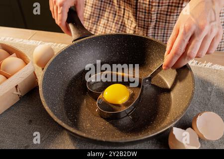 Nahaufnahme von weiblichen Händen, die eine Herzform auf einer Pfanne halten, mit einem gebrochenen Ei, herzförmigem Spiegelei Stockfoto