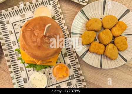 Ein großer, mehrschichtiger Burger mit panierten Hähnchenfilets, Gemüse, viel Käse und Saucen neben einem Teller panierter Nuggets Stockfoto