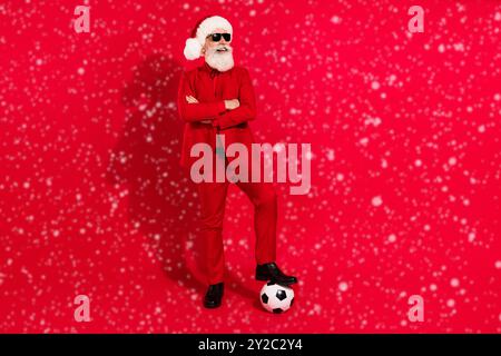 Foto von lustigen coolen Mann gekleidet weihnachten Kostüm Hut Arme gefaltet spielen Fußball leeren Raum isoliert roten Hintergrund Stockfoto