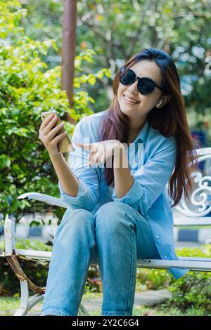 Eine reife indonesische Frau mit Sonnenbrille schaut fröhlich in die Kamera und zeigt auf ein Handygerät, das in einem Park im Freien sitzt. Studentinnen für Stockfoto