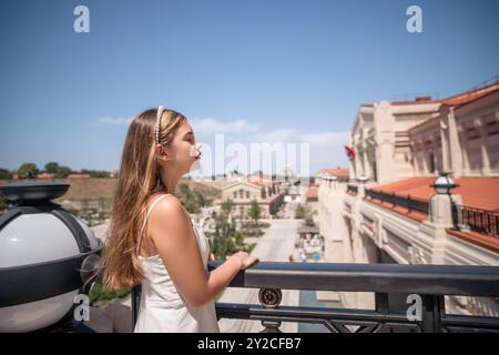 Eine Frau in einem weißen Kleid steht auf einem Balkon und blickt auf ein Stadtbild unter einem klaren blauen Himmel. Stockfoto