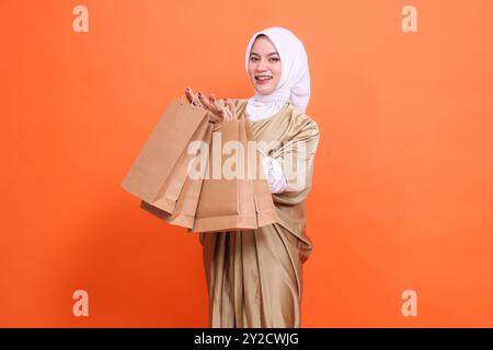 Fröhliche asiatische Frau mit beiden Händen, die eine braune Papiertüte mit Lebensmitteln vor sich trägt, in einem muslimischen Kaftan-Kleid mit einem Hijab, isola Stockfoto
