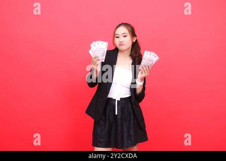 Geste einer jungen indonesischen Bürofrau, die die Stirn trägt, in einem schwarzen formalen Anzug, beide Hände mit Rupiah-Geld, für finanzielle Inhalte, Geschäfte, Werbung Stockfoto